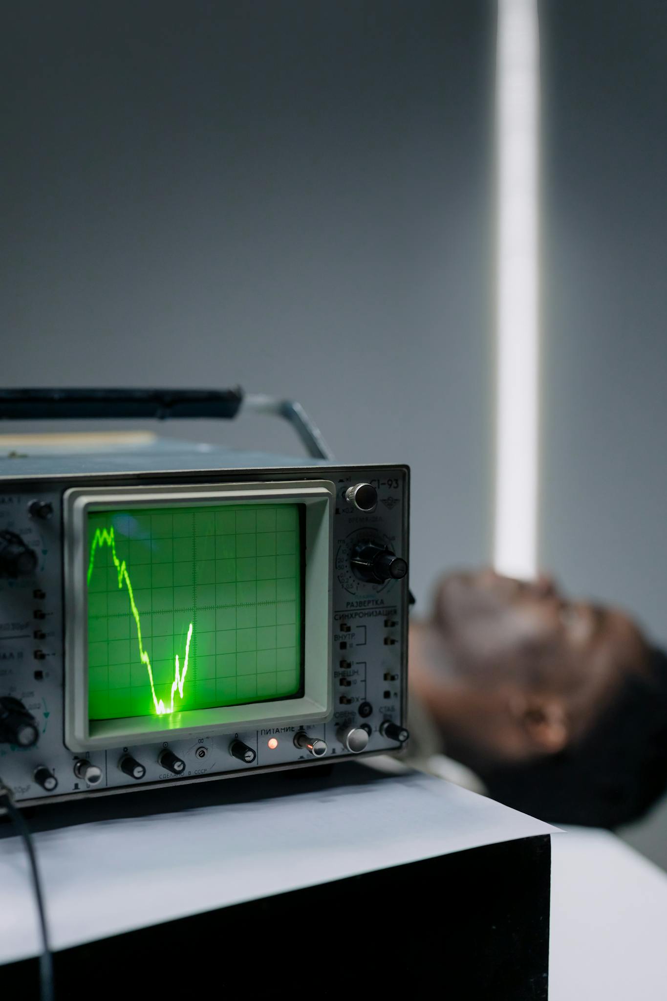 Close-up of an oscilloscope displaying a pulse signal beside a resting man indoors.